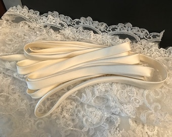 Op de afbeelding: Een opgerolde lengte glad, gebroken wit satijnen lint dat op een stuk witte kanten stof met bloemmotief ligt. Het lint heeft een glanzende afwerking en is in meerdere lussen gevouwen, wat contrasteert met het ingewikkelde, opengewerkte patroon van het kant.