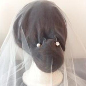 May include: A sheer white bridal veil draped over a dark-haired mannequin head. The veil is secured with two decorative hair pins, one featuring a round white pearl and the other a clear, faceted crystal stone, positioned on the back of the head.