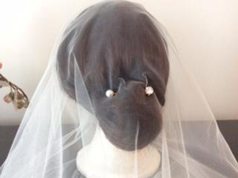 May include: A sheer white bridal veil draped over a dark-haired mannequin head. The veil is secured with two decorative hair pins, one featuring a round white pearl and the other a clear, faceted crystal stone, positioned on the back of the head.
