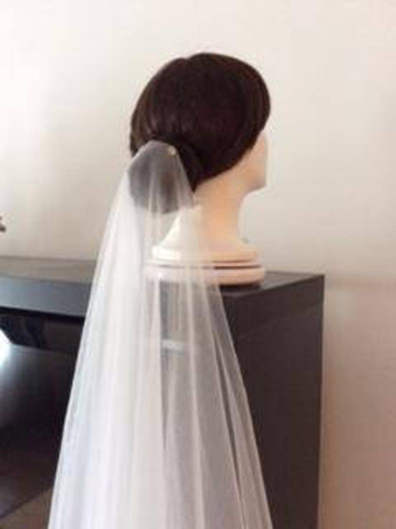 May include: A white bridal veil made of tulle, with a single pearl embellishment at the top. The veil is draped over a mannequin head, with the veil flowing down the back.