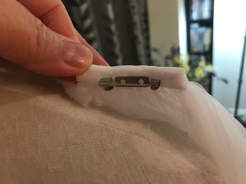 May include: A white tulle fabric with a silver safety pin attached to it.