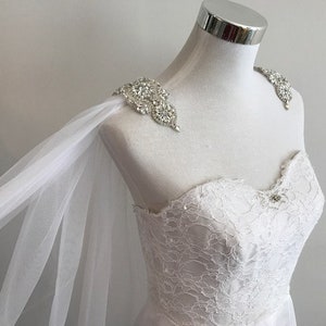 May include: White tulle bridal cape with crystal embellishments on the shoulders. The cape is draped over a white lace wedding dress.