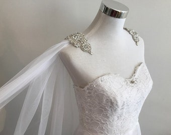 May include: White tulle bridal cape with crystal embellishments on the shoulders. The cape is draped over a white lace wedding dress.