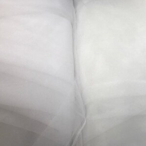 May include: A folded length of sheer white tulle fabric with a fine mesh texture. The material is translucent and lightweight, displaying soft folds and layers that create subtle variations in opacity across the surface of the textile.