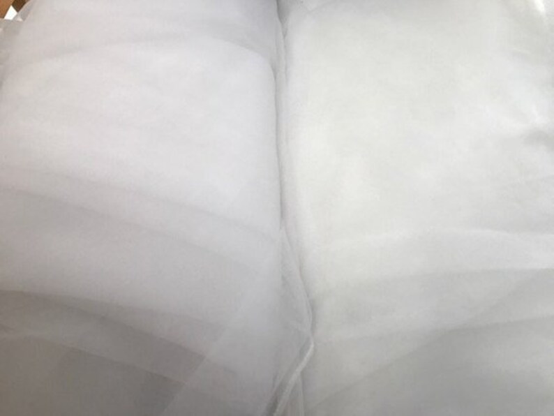 May include: A folded length of sheer white tulle fabric with a fine mesh texture. The material is translucent and lightweight, displaying soft folds and layers that create subtle variations in opacity across the surface of the textile.