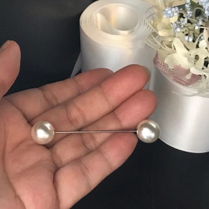 May include: A straight metal pin featuring two smooth, round, white faux pearls at each end. The pin is thin and metallic, with one pearl positioned on the left and one on the right, creating a balanced, linear design for garment fastening.