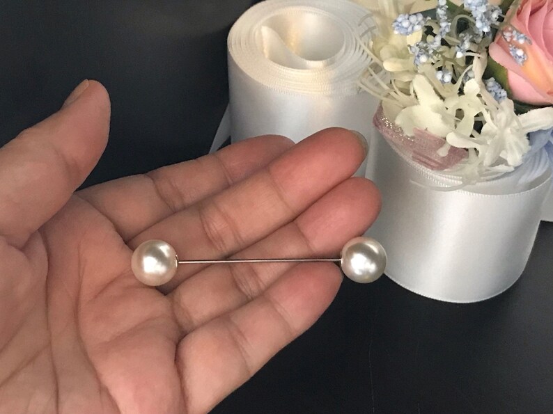 May include: A straight metal pin featuring two smooth, round, white faux pearls at each end. The pin is thin and metallic, with one pearl positioned on the left and one on the right, creating a balanced, linear design for garment fastening.