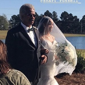 Op de afbeelding: Een bruid in een witte trouwjurk loopt met haar vader de gang door. Ze draagt ​​een sluier en draagt ​​een boeket witte bloemen. De ceremonie vindt buiten plaats, met een meer op de achtergrond.