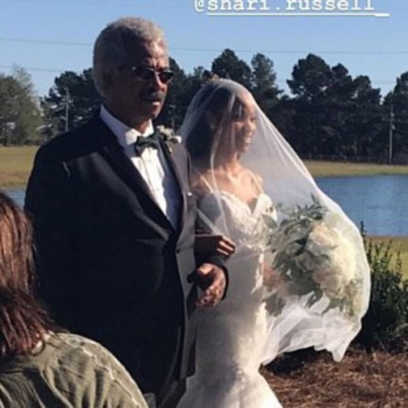 Op de afbeelding: Een bruid in een witte trouwjurk loopt met haar vader de gang door. Ze draagt ​​een sluier en draagt ​​een boeket witte bloemen. De ceremonie vindt buiten plaats, met een meer op de achtergrond.