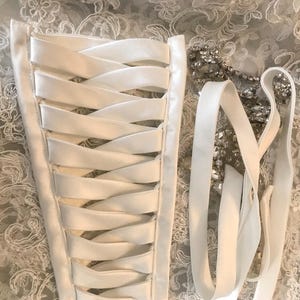 May include: A white satin corset back panel featuring a crisscross ribbon lacing system. Beside the panel lies a long white satin ribbon and a decorative strand of clear, faceted crystals, all set against a white floral lace fabric.