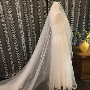 Op de afbeelding: Een witte tule bruids sluier met een lange sleep. De sluier is gedrapeerd over een witte jurk en wordt getoond op een mannequin.