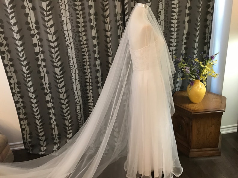 Op de afbeelding: Een witte tule bruids sluier met een lange sleep. De sluier is gedrapeerd over een witte jurk en wordt getoond op een mannequin.