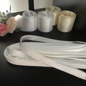 May include: A collection of satin ribbon rolls in white and cream shades, accompanied by a loose, folded length of white satin ribbon. A small floral arrangement featuring pink, white, and blue artificial flowers is positioned to the left.