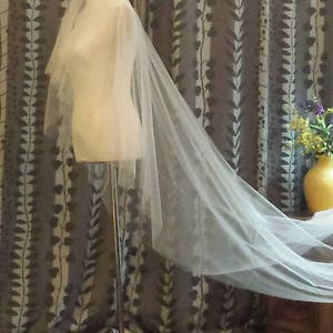 May include: A long, sheer, off-white bridal veil draped over a mannequin torso. The veil features multiple layers of fine, translucent tulle fabric that extend downward and trail behind the form, creating a soft, flowing, and elegant appearance.