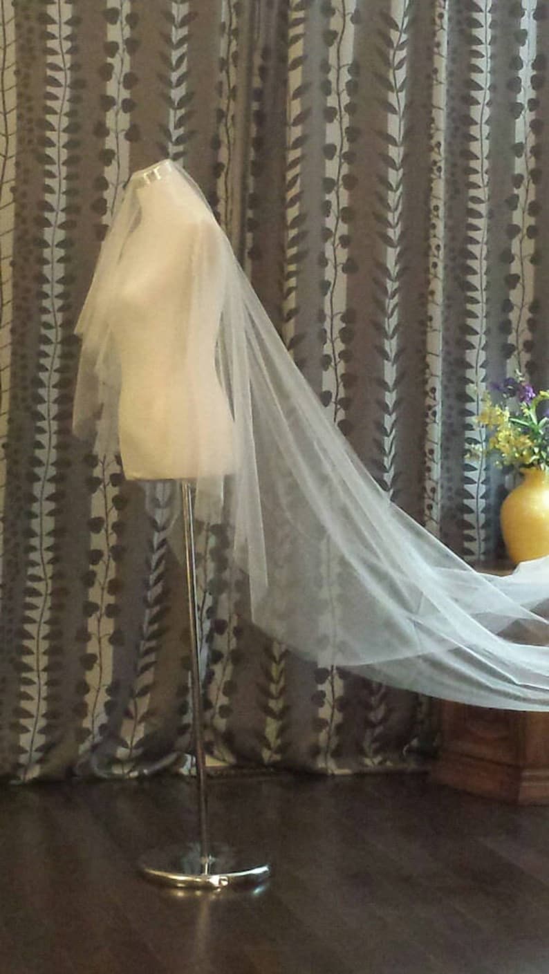 May include: A long, sheer, off-white bridal veil draped over a mannequin torso. The veil features multiple layers of fine, translucent tulle fabric that extend downward and trail behind the form, creating a soft, flowing, and elegant appearance.