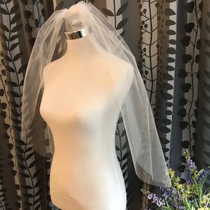 May include: A sheer white bridal veil with a silver band is displayed on a mannequin. The veil is draped over the mannequin's head and shoulders, with a long, flowing design. The background features a patterned curtain.