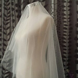 May include: A sheer, off-white bridal veil made of fine tulle fabric draped over a mannequin. The veil features a smooth, lightweight texture and a simple, unadorned edge, cascading down from the top of the mannequin to create a soft, layered effect.