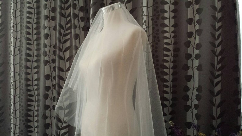 May include: A sheer, off-white bridal veil made of fine tulle fabric draped over a mannequin. The veil features a smooth, lightweight texture and a simple, unadorned edge, cascading down from the top of the mannequin to create a soft, layered effect.