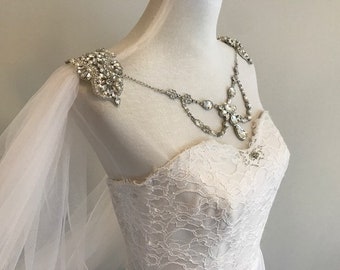 Op de afbeelding: Een witte bruidsjurk met een kantboord en een tule overlay. De jurk heeft een ketting van kristallen en strass en een bijpassend schouderstuk.