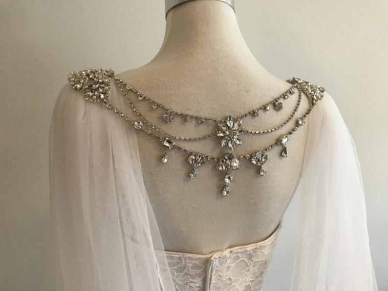 May include: A white tulle bridal cape with a crystal and rhinestone embellishment. The embellishment features a delicate chain design with dangling crystals and a floral motif.