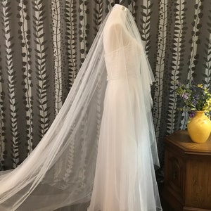 May include: A white bridal veil made of tulle with a long train. The veil is draped over a white dress and is displayed against a patterned background.