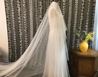 May include: A white bridal veil made of tulle with a long train. The veil is draped over a white dress and is displayed against a patterned background.