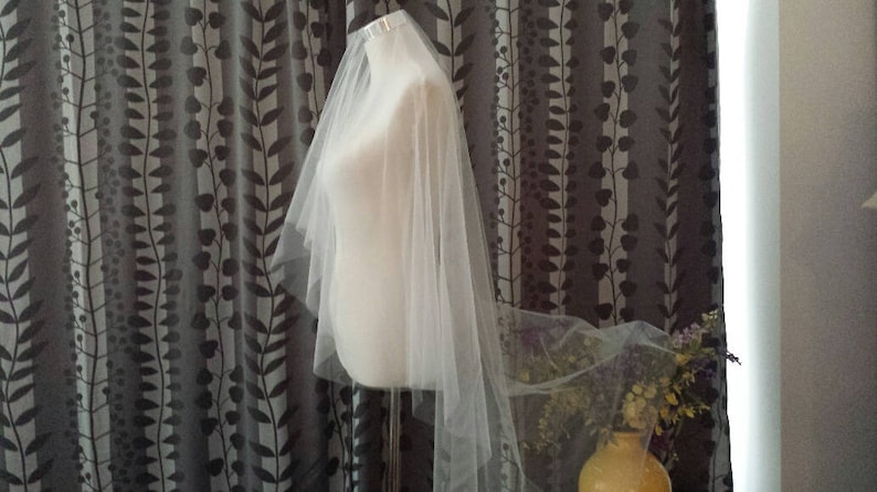 May include: A white tulle wedding veil with a single layer and a scalloped edge. The veil is displayed on a mannequin against a patterned background.