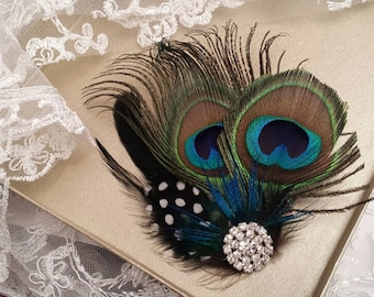 May include: A decorative hair accessory featuring two iridescent peacock feathers with blue and green eye patterns, layered over black and white polka-dot feathers, and accented with a round, clear rhinestone-encrusted silver-tone metal brooch.