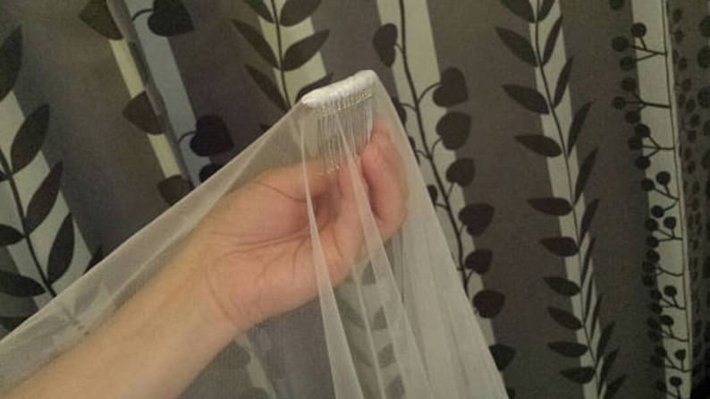 May include: A white bridal veil made of sheer, lightweight tulle fabric. The top of the veil is gathered and securely attached to a silver-toned metal hair comb with multiple teeth, designed to be inserted into hair for a secure hold.