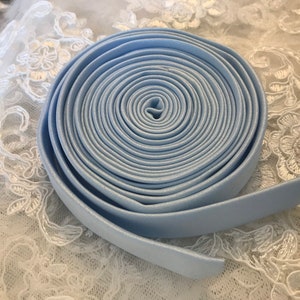 May include: A tightly coiled roll of light blue satin ribbon. The ribbon is smooth and has a slight sheen, with a visible edge. The ribbon is displayed on a white lace fabric, creating a soft contrast.