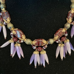 May include: A gold and purple beaded necklace with a decorative design. The necklace features a series of oval-shaped beads with a striped pattern in shades of brown and purple. The beads are interspersed with smaller gold beads and decorative gold accents. The necklace is finished with a series of purple tassel-like pendants.