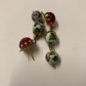 May include: A pair of dangle earrings featuring gold-tone metal settings, red carved glass beads, and white spherical beads decorated with multi-colored floral patterns in green, red, and brown, connected by small gold-tone metal links and caps.