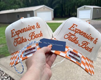 Peut inclure: Deux casquettes de baseball blanc cassé avec le texte brodé en orange Expensive & Talks Back. Les visières présentent un motif à carreaux orange et blanc. Une étiquette bleue marquée The Triune Creative est attachée par un cordon bleu et blanc.