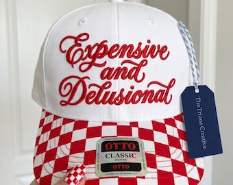 Peut inclure: Une casquette de baseball blanche avec le texte brodé en rouge Expensive and Delusional sur le devant. La visière présente un motif à carreaux rouges et blancs. Une étiquette bleue marquée The Triune Creative est attachée par un cordon rayé.