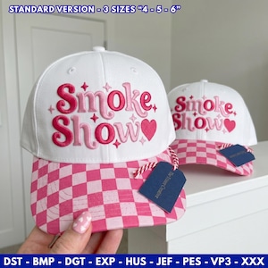 May include: Two white baseball caps with pink checkered brims. The front of each cap features the embroidered pink text Smoke Show with small star accents and a heart. A dark blue tag is attached to the brim with a red and white string.