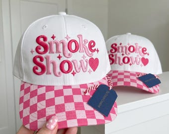 Peut inclure: Deux casquettes de baseball blanches avec des visières à carreaux roses. L'avant de chaque casquette présente le texte rose brodé Smoke Show avec des petites étoiles et un cœur. Une étiquette bleu foncé est attachée à la visière.