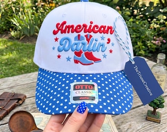 Peut inclure: Une casquette de baseball blanche avec une visière à pois bleus. L'avant présente des bottes de cow-boy brodées en rouge et bleu avec le texte "American Darlin" en écriture rouge et bleue. Une étiquette bleue "TheTriuneCreative" est attachée.
