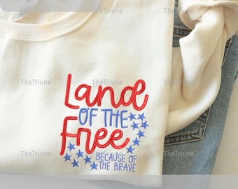 Peut inclure: Sweat-shirt crème avec la phrase brodée "Land of the Free" en rouge et bleu, entourée d'étoiles bleues. Le texte "Because of the Brave" est en dessous. L'image affiche également les tailles 12.7cm, 15.2cm, 17.8cm et 20.3cm.
