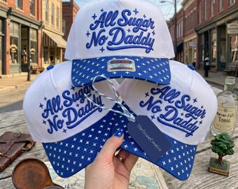 Peut inclure: Trois casquettes de baseball blanches avec une broderie bleue indiquant All Sugar No Daddy et de petites étoiles. Les visières présentent un motif de croix bleu et blanc. Une étiquette bleu foncé marquée TheTriuneCreative est attachée.
