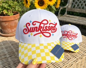 Peut inclure: Deux casquettes de baseball blanches avec des visières à carreaux jaunes et blancs. L'avant de chaque casquette présente le texte brodé en rouge Sunkissed dans une police cursive, agrémenté de petites étoiles brodées bleues et jaunes.