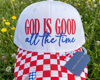 Peut inclure: Une casquette de baseball blanche avec le texte brodé en rouge GOD IS GOOD et le texte en cursive bleue all the time. La visière présente un motif à carreaux rouges et blancs. Une étiquette bleue marquée The Triune Creative est attachée.