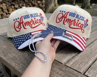 Peut inclure: Deux casquettes de baseball blanc cassé avec le texte brodé en rouge et bleu "GOD BLESS America" et des étoiles. Les visières présentent un motif patriotique, l'une avec des étoiles blanches sur fond bleu et l'autre avec des rayures rouges.