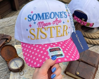 Peut inclure: Une casquette de baseball blanche avec une visière à motif floral rose et le texte brodé SOMEONE'S Mean Ass SISTER en bleu, rose et doré. La casquette présente un bouton rose sur le dessus, une étiquette rayée bleu et blanc et une étiquette bleue.