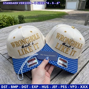 May include: Two cream-colored baseball caps with blue and white polka dot brims. The front panels feature gold embroidered text reading WRONGHOLE but i LIKE IT. A small blue tag labeled The Triune Creative is attached to the caps.