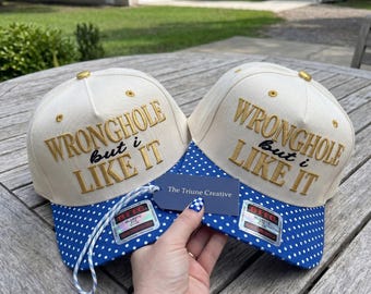 Peut inclure: Deux casquettes de baseball couleur crème avec des visières à pois bleus et blancs. Les panneaux avant présentent le texte brodé en doré WRONGHOLE but i LIKE IT. Une petite étiquette bleue marquée The Triune Creative est fixée aux casquettes.