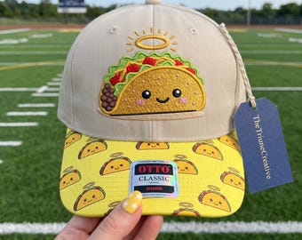 Peut inclure: Une casquette de baseball beige avec un taco brodé surmonté d'une auréole sur le panneau avant. La visière jaune présente un motif répétitif de petits tacos avec des auréoles. Une étiquette bleu foncé marquée TheTriuneCreative est attachée.