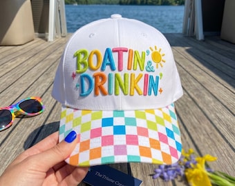 Peut inclure: Une casquette de baseball blanche avec le texte brodé "BOATIN & DRINKIN" en lettres multicolores, une icône de soleil jaune et un motif à carreaux colorés sur la visière. Le texte "STANDARD VERSION - 3 SIZES - 4 - 5 - 6" est visible en haut.