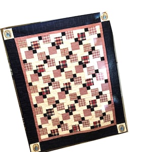 May include: A quilt with a patchwork design featuring red, black, and white plaid squares on a cream background. The quilt is bordered with a dark blue fabric and a pink trim.