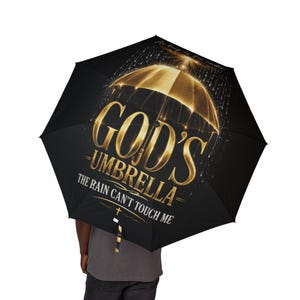 May include: Black umbrella with gold text that reads "GOD'S UMBRELLA - THE RAIN CAN'T TOUCH ME." The umbrella has a gold illustration of an umbrella with rain falling around it. The umbrella is held by a person wearing a gray shirt.