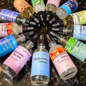 May include: A circular arrangement of twelve clear glass dropper bottles containing Hyaluronic Serum. Each bottle features a black dropper cap and a label with specific ingredients like Niacinamide, Frankincense, Collagen, and Calendula, each holding 2 fl oz.